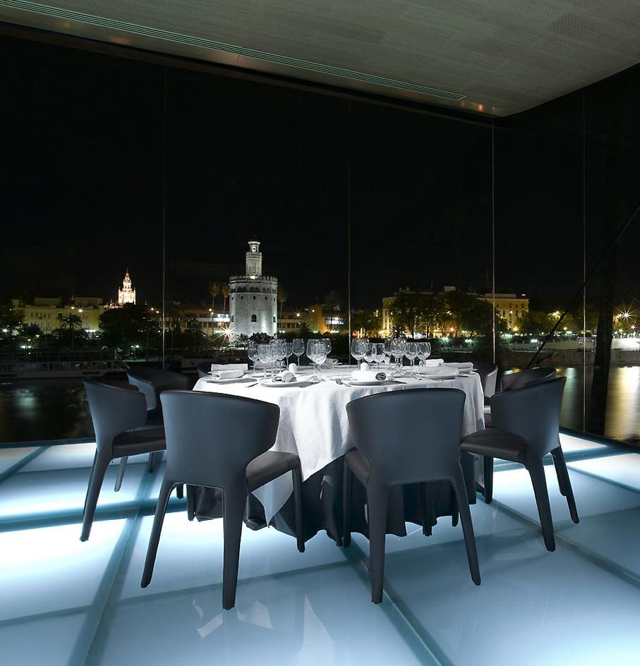 Mesa redonda de cena en un salón acristalado con la Torre del Oro iluminada al fondo